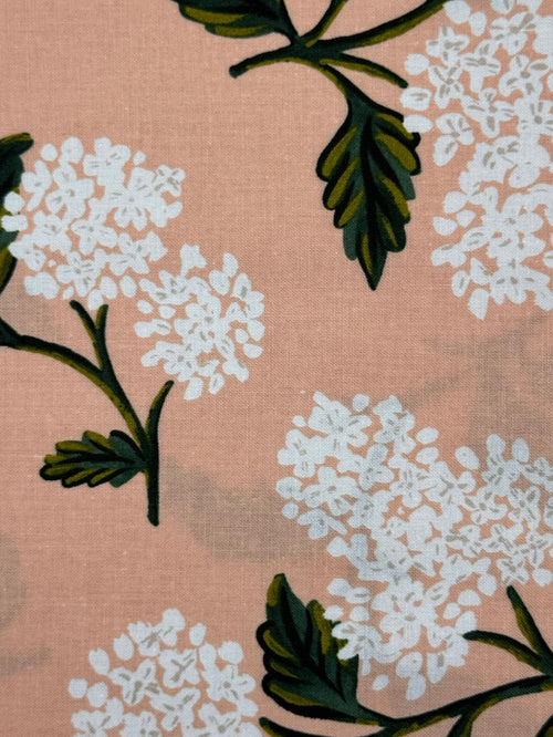 A close-up of a fabric pattern featuring white flower clusters and dark green leaves on a dusty pink background.