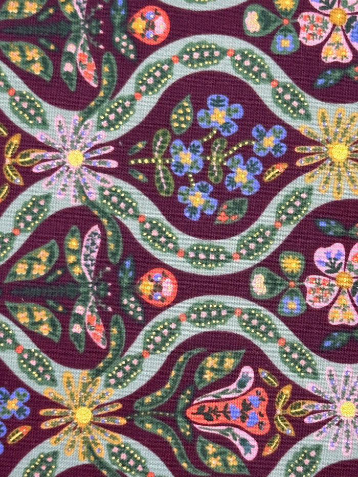 A close-up of a patterned fabric featuring a dark purple background with a repeating folk-art style design of green leaves, blue flowers, pink blossoms, and yellow sunflowers.