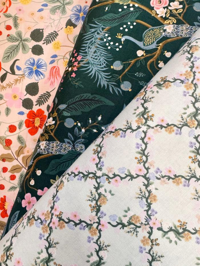 Three folded fabric samples with distinct floral and nature prints layered diagonally. The top-left fabric is light pink with red, blue, and green flowers.