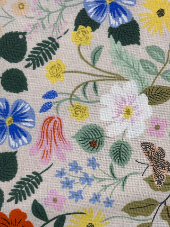 A close-up of a fabric pattern featuring a variety of colorful wildflowers, green leaves, and a butterfly on a light beige linen-like background.