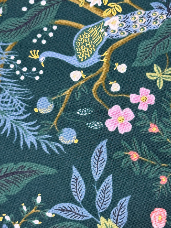 A close-up of a fabric pattern featuring a peacock, pink flowers, and botanical leaves on a dark teal background.