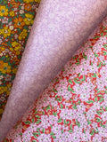 Three rolls of patterned fabric stacked diagonally, showing different designs: a yellow and orange floral print, a light lavender floral print, and a red floral print with pink blossoms and green leaves.