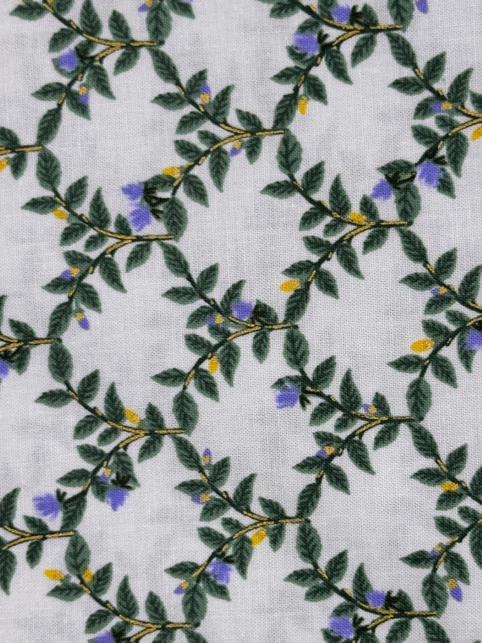A close-up of a white textured fabric featuring a repeating botanical pattern of green leaves, thin brown branches, and small purple and yellow flowers.