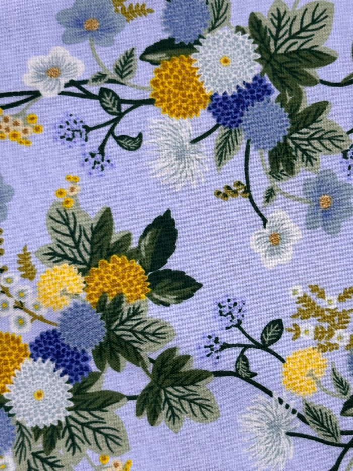 A close-up of a floral fabric pattern on a light lavender background, featuring white, blue, and yellow flowers with green leaves and stems.